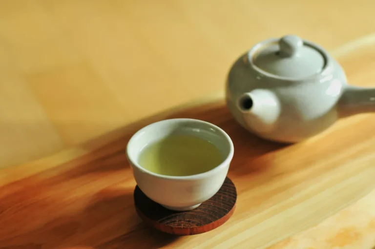A ceramic teacup filled with light green tea sits on a wooden coaster, next to a matching teapot on a smooth, light wood surface. The scene feels calm and serene.