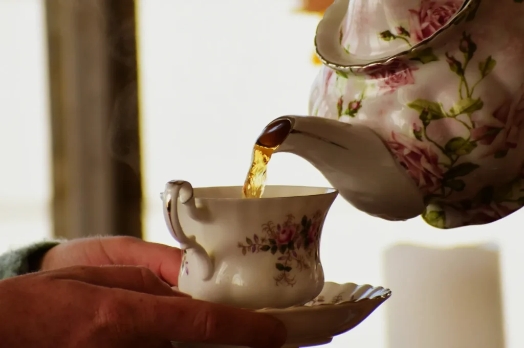 A floral teapot pours steaming tea into a matching cup held by a hand, evoking warmth and elegance in a cozy, intimate setting.