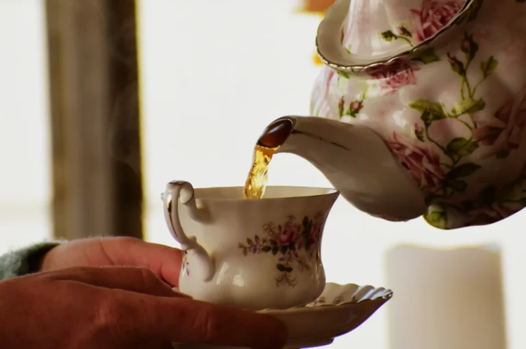 A floral teapot pours steaming tea into a matching cup held by a hand, evoking warmth and elegance in a cozy, intimate setting.