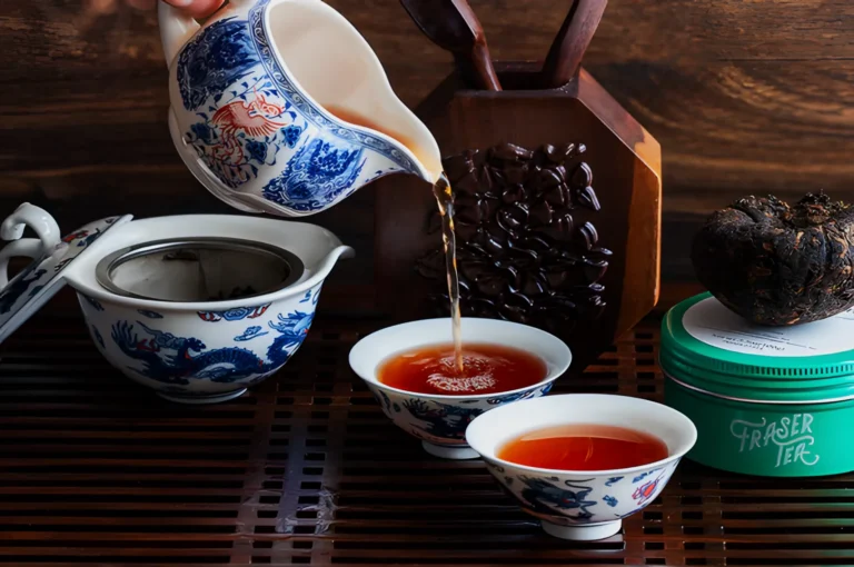 A hand pours tea from a blue-and-white porcelain teapot into matching cups on a wooden tray. A green container and a tea brick are in the background.