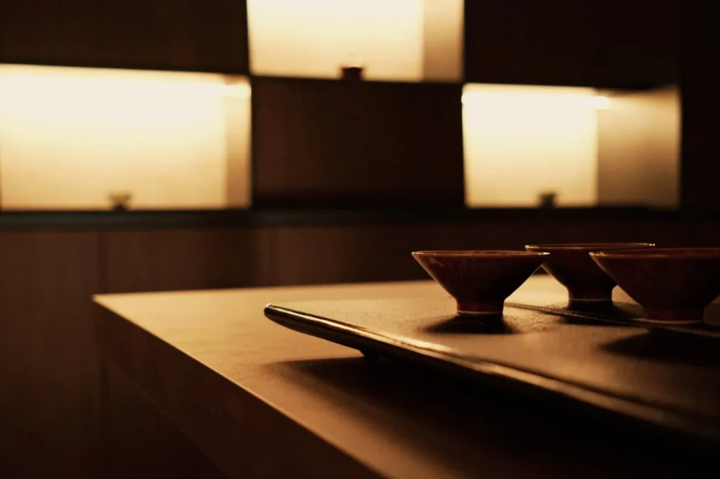 A minimalist interior features softly lit shelves and a wooden countertop with a black tray holding small, elegant cups. The scene feels warm and serene.