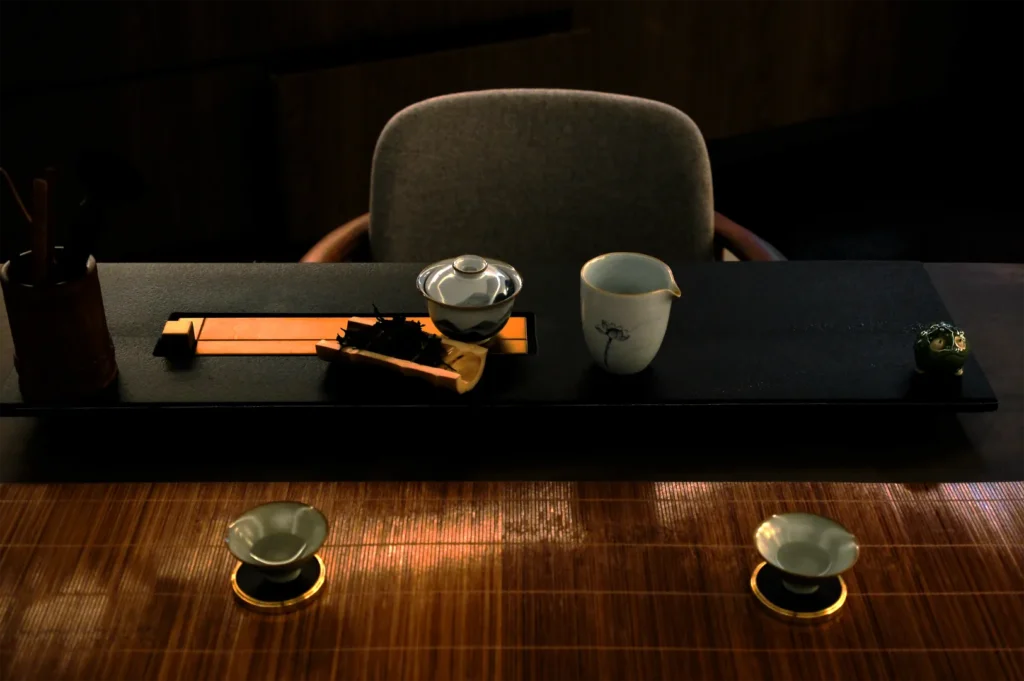 Dimly lit tea ceremony setup with a modern teapot, cup, and utensils on a dark tray. Two small bowls with saucers sit on a bamboo mat, evoking a serene atmosphere.