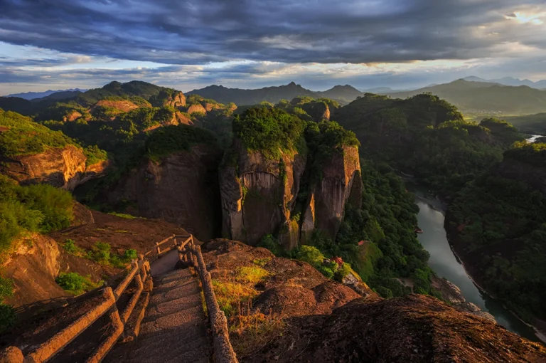 Stone steps lead to a viewpoint overlooking lush, rugged mountains and a winding river under a dramatic, cloudy sky. The scene feels serene and majestic.