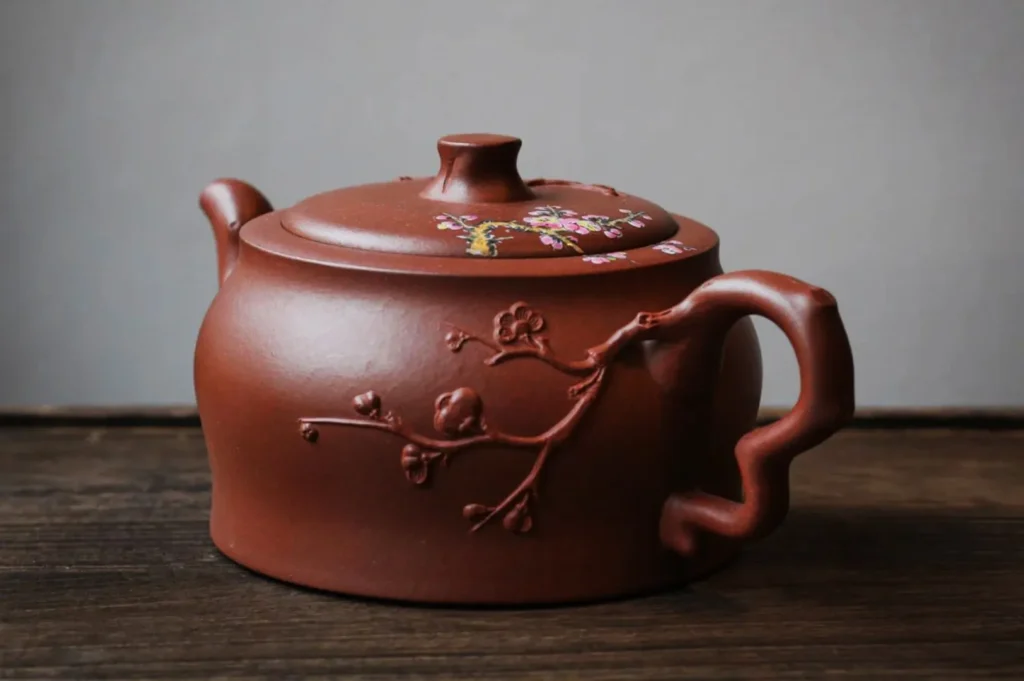 A clay teapot with a cherry blossom branch design and subtle pink and yellow accents on the lid, set on a wooden surface. Elegant and traditional.