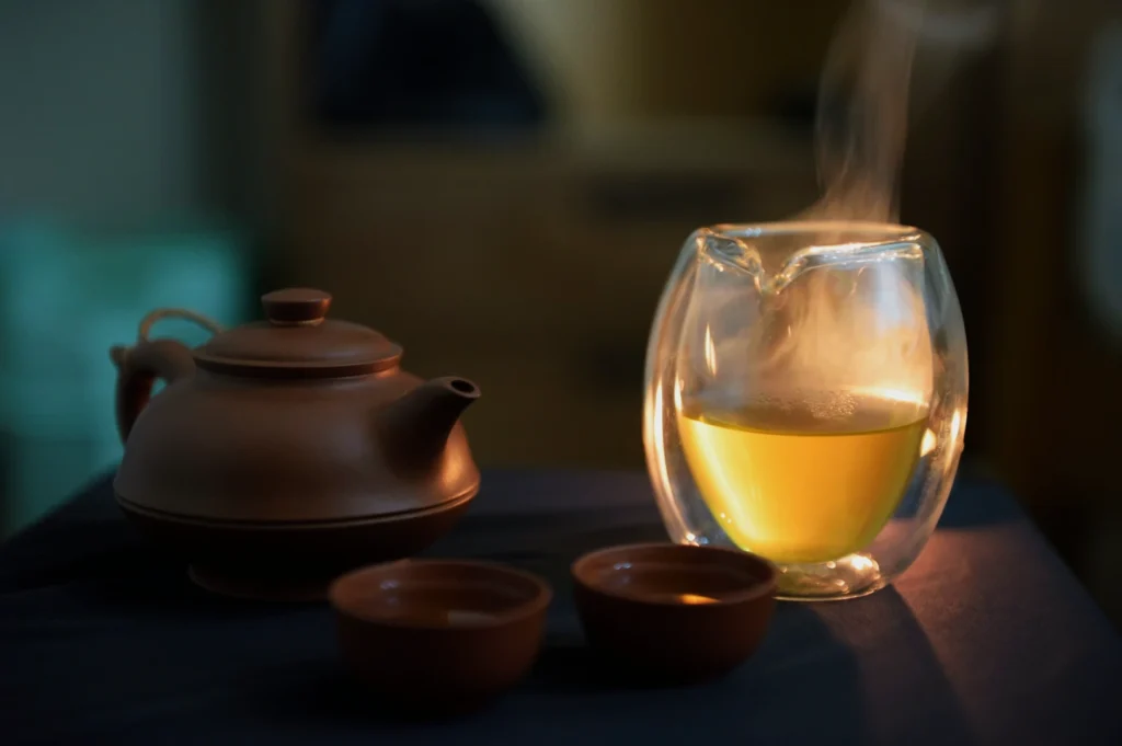 Steaming glass cup of tea, next to a brown teapot and two small cups, creates a warm, inviting mood on a dimly lit table.