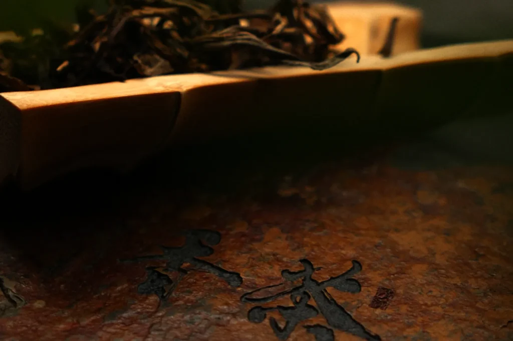A wooden tray with dried tea leaves rests on a textured, reddish-brown surface. Chinese characters are engraved below, creating a calm, rustic ambiance.