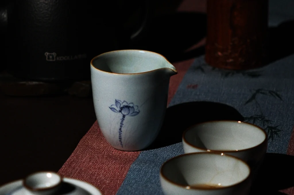 A soft-lit ceramic jug with a delicate lotus design on a blue and red fabric. Nearby, two small matching cups create a calm, elegant tea setting.