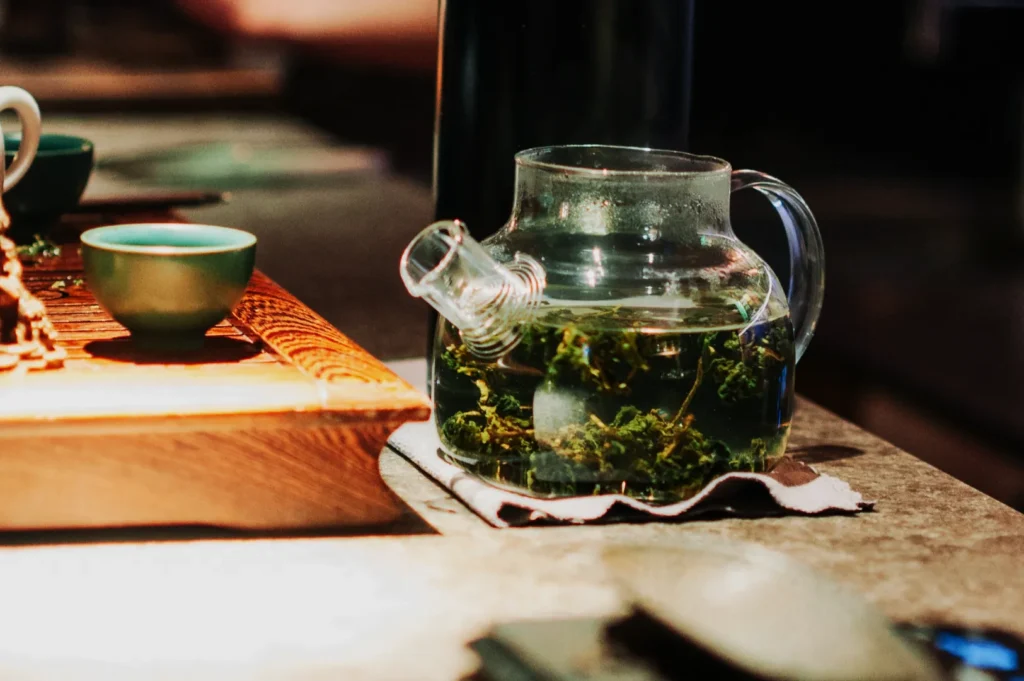 Clear glass teapot with loose green tea steeping inside, placed on a table beside a wooden tray with a small teal cup, in a cozy setting.