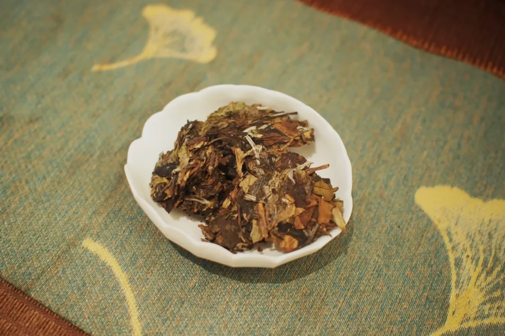 A white bowl filled with loose, dried tea leaves sits on a teal-patterned fabric with yellow floral designs, creating a calm, rustic atmosphere.