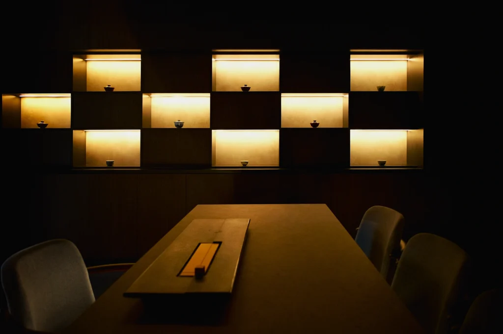 Dimly lit room with illuminated display shelves, each holding a small bowl. A long table and chairs suggest a minimalist, serene atmosphere.