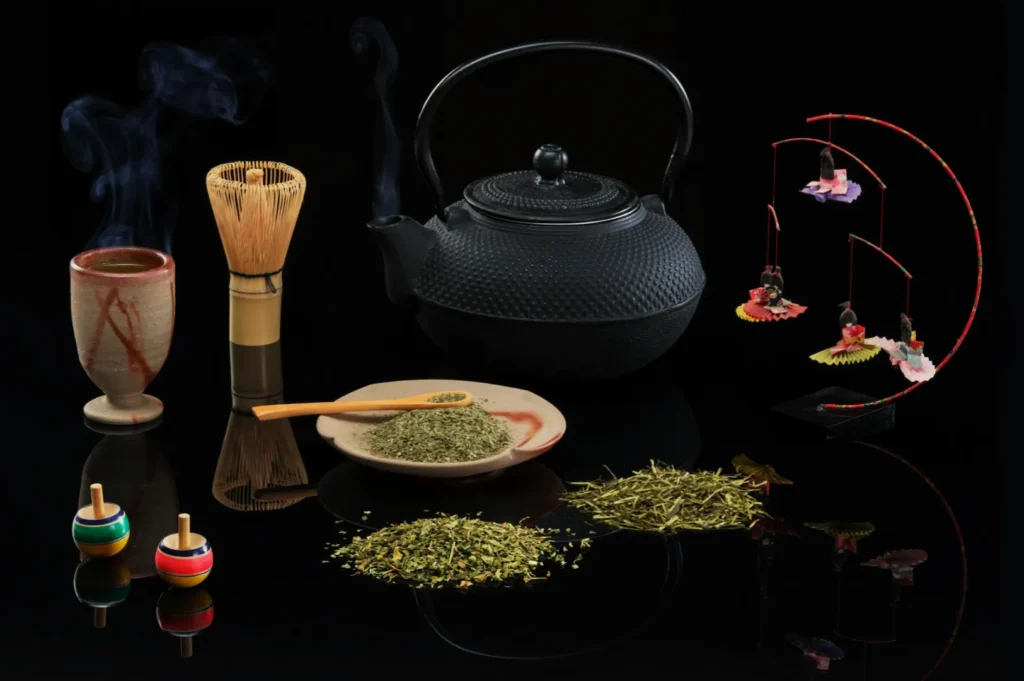 Japanese tea ceremony setup with a black teapot, steaming cup, bamboo whisk, green tea on a plate, colorful spinning tops, and hanging ornaments.