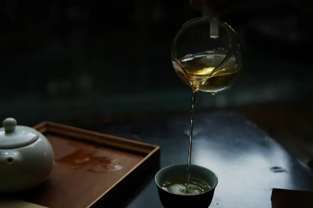 A hand pours clear, light-colored tea from a glass pitcher into a small cup on a dark table. A white teapot and wooden tray are nearby, creating a calm, cozy atmosphere.
