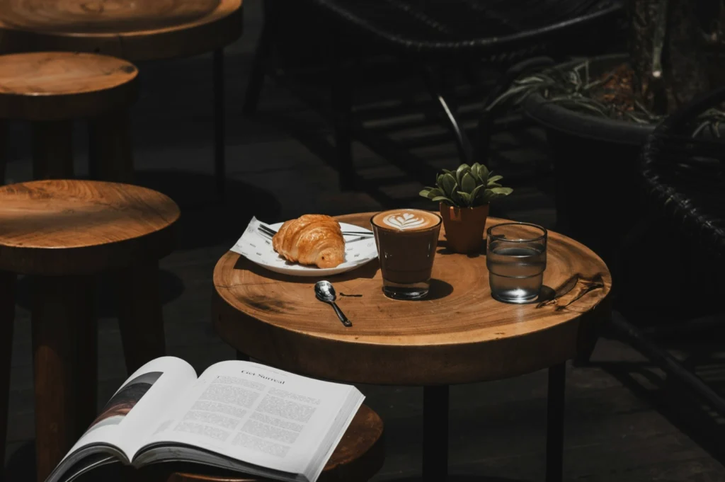 Wooden table with a croissant on a plate, a cappuccino with latte art, a glass of water, a spoon, a small potted plant, and an open book, creating a cozy cafe atmosphere.