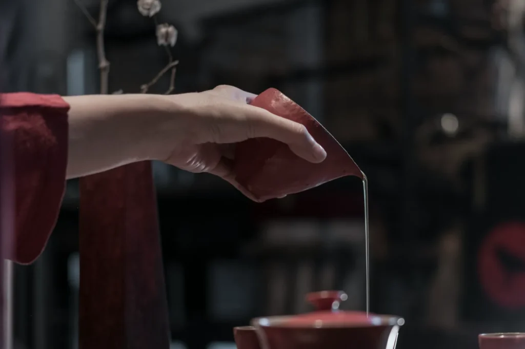 Hand in a red sleeve pours liquid from a tilted red pitcher into a red pot. Soft lighting and blurred background create a serene atmosphere.