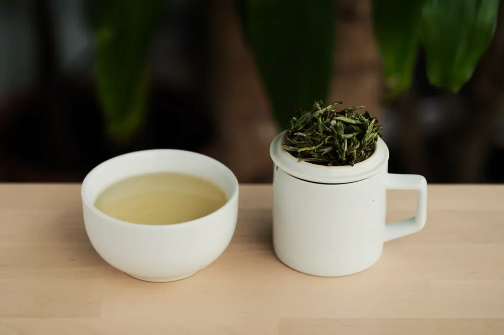White ceramic cup filled with light green tea next to a mug with a lid topped with loose tea leaves. Set on a wooden table with plants blurred in the background.