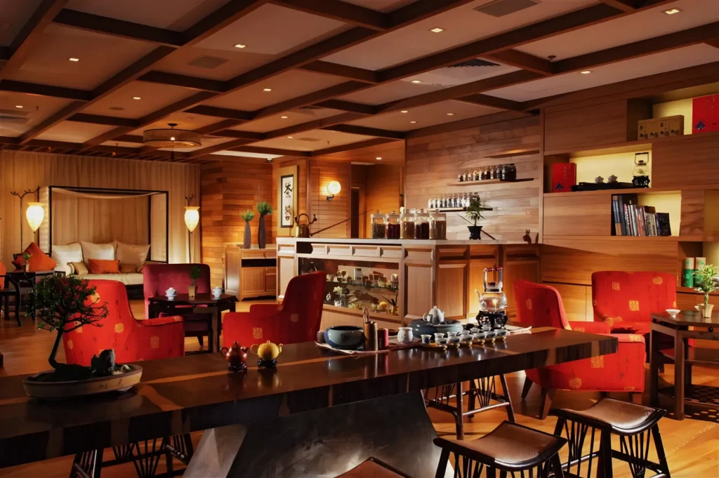 Elegant tea room with warm lighting and wooden decor. Red chairs and tea sets on tables evoke a cozy, inviting atmosphere. Indoor plants add freshness.