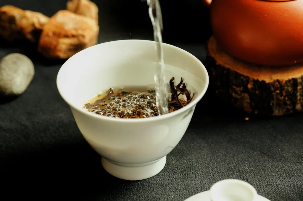 Water is being poured into a white cup filled with tea leaves, creating bubbles. There's a clay teapot on a wooden stand, with stones in the background.