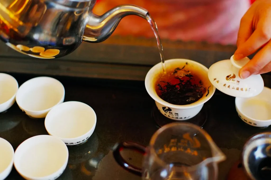 A person pours hot water from a metal kettle into a gaiwan filled with tea leaves on a tray. Empty white cups are arranged nearby, creating a serene, focused atmosphere.