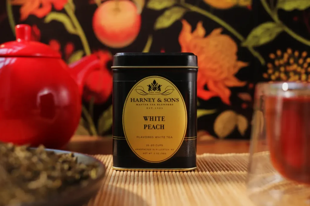 A tin of Harney & Sons White Peach tea on a mat, flanked by a red teapot and a glass of tea. Floral wallpaper creates a warm, inviting backdrop.
