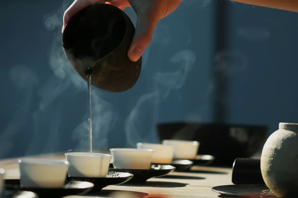 A hand pours steaming tea from a rustic teapot into small white cups on a sunlit table, creating a peaceful and serene atmosphere.