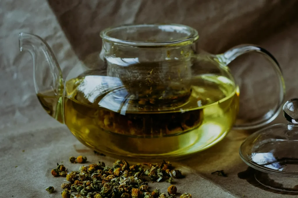 Glass teapot with chamomile tea, surrounded by loose chamomile flowers, sits on a textured surface. The scene feels calm and inviting.