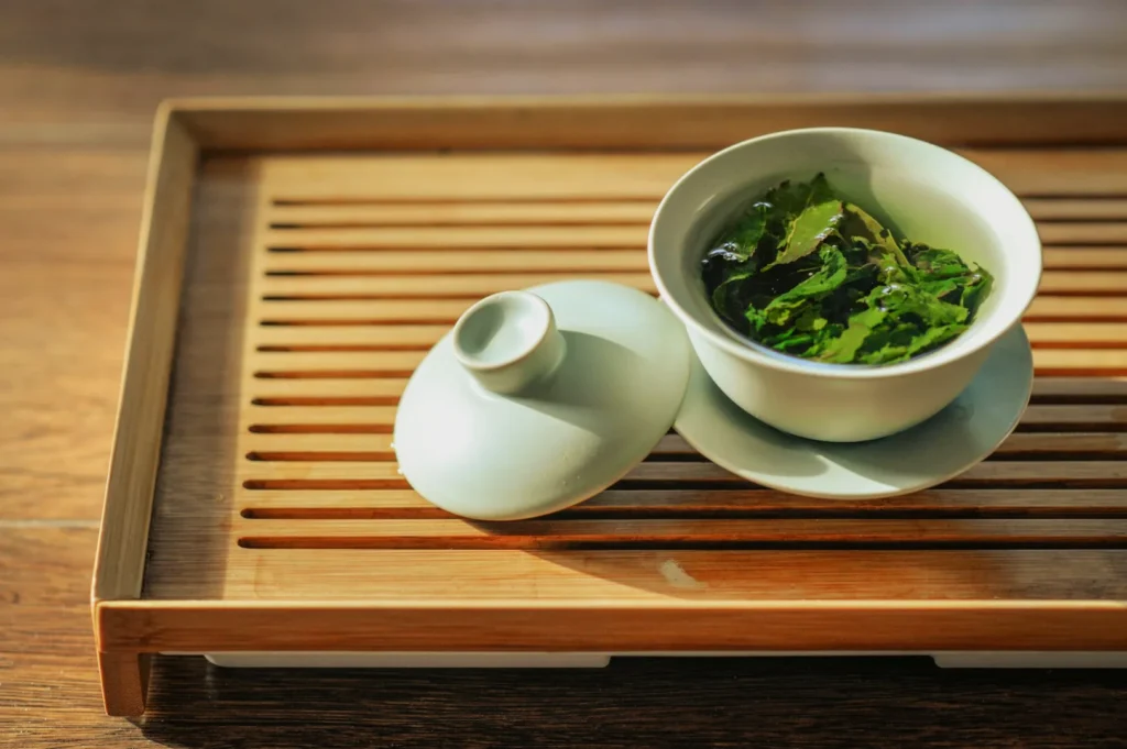A serene tea scene featuring a traditional Chinese gaiwan with a lid, filled with steeping green tea leaves on a slatted bamboo tray, softly lit by the sun.