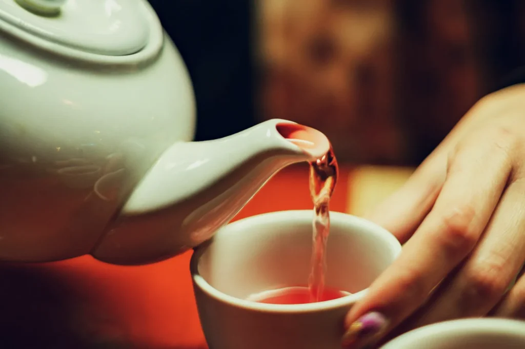 A white teapot pours red tea into a small cup, held by a hand with painted nails. The warm lighting creates a cozy and inviting atmosphere.