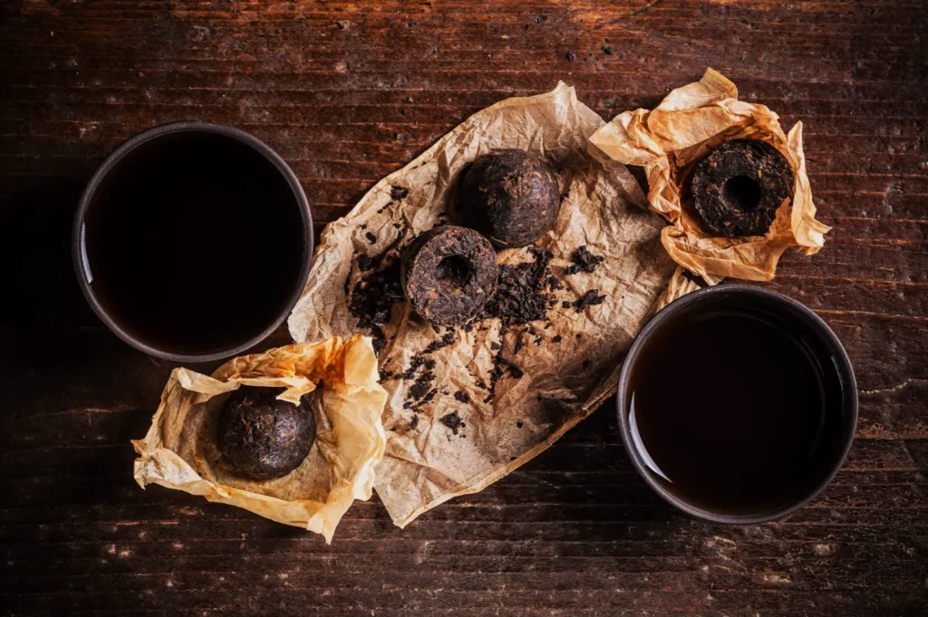 Three dark, round tea balls sit on crumpled paper, surrounded by loose tea leaves. Two cups of dark tea are nearby on a rustic wooden table.