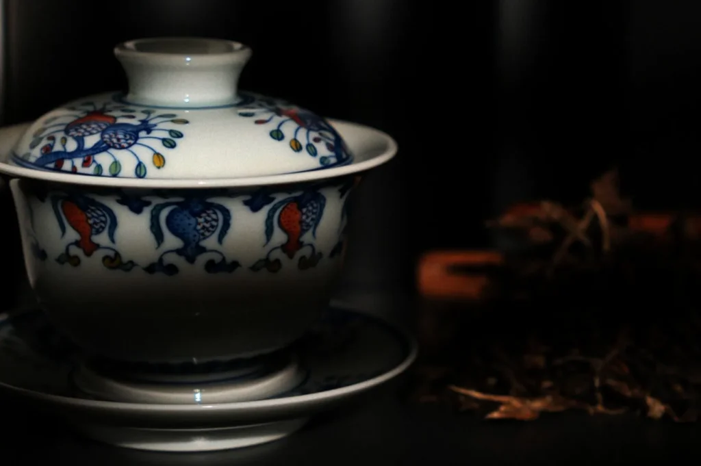 A white porcelain tea cup with a lid and saucer, adorned with colorful blue and red peacock motifs, sits elegantly beside loose tea leaves.