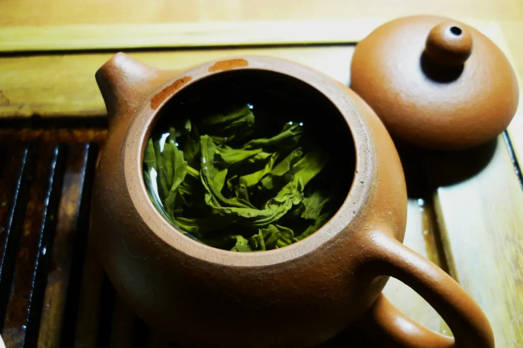 A clay teapot with its lid resting beside it, revealing green tea leaves steeping in water, set on a wooden tray. The scene feels warm and inviting.