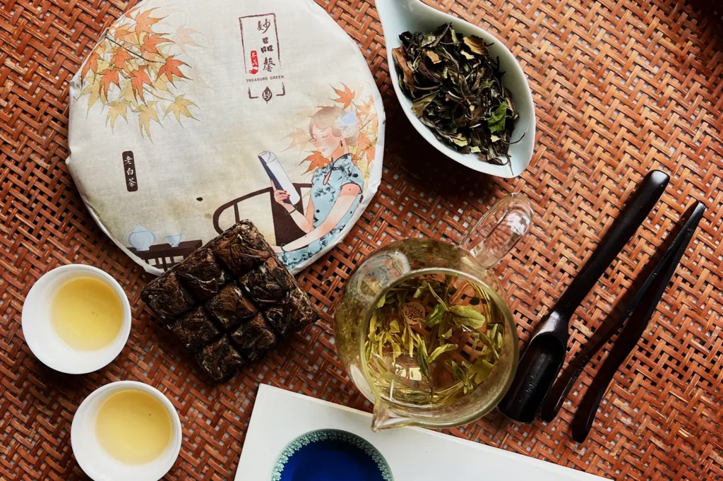 A flat lay of a tea setup on a woven mat. Includes a wrapped tea cake, teapot, teacups, dried leaves in a dish, tea picks, and a blue dish. Warm and inviting.