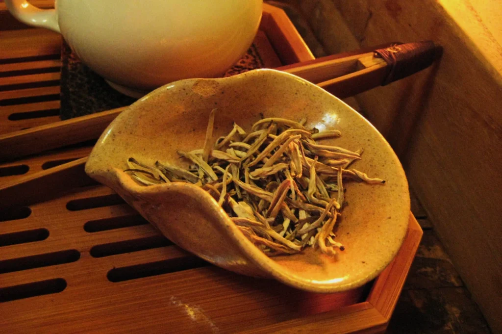 A ceramic dish holds dried silver needle tea leaves on a wooden tea tray. The warm lighting creates a cozy and inviting atmosphere.