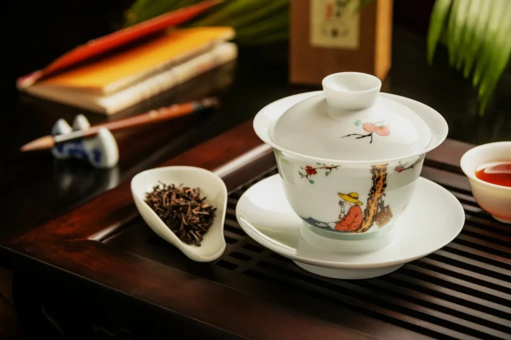 Elegant ceramic gaiwan with intricate design on a wooden tray, surrounded by a dish of loose tea leaves, a cup of tea, books, and chopsticks, creating a serene tea ceremony ambiance.