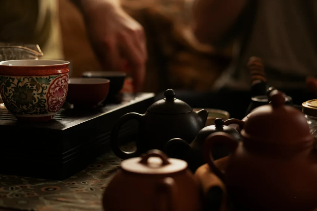 A cozy tea setup features ornate teacups and several clay teapots on a wooden tray. Hands are visible, adding a sense of calm and gathering.