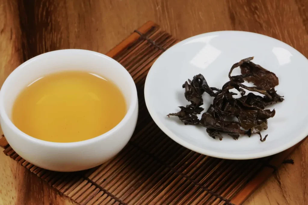 A white cup of golden oolong tea and a white saucer with loose tea leaves sit on a bamboo mat, evoking a calm, traditional tea ritual.