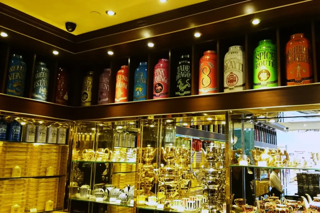 A row of colorful, large tea canisters on a shelf above a display of shiny, golden teapots and cups. The setting conveys a luxurious, elegant atmosphere.