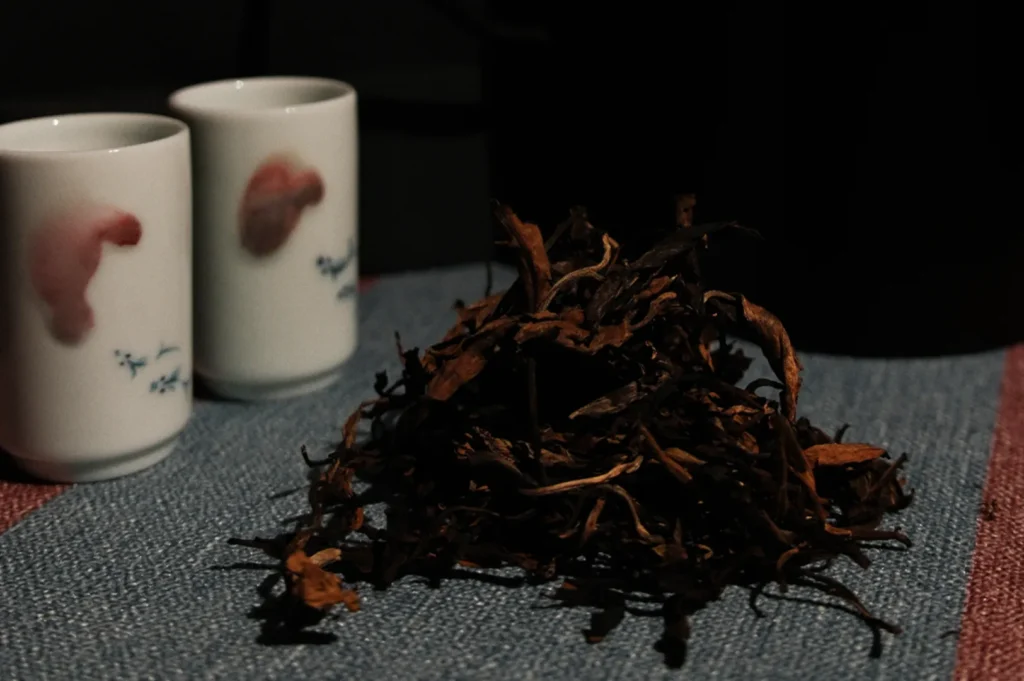 A pile of dried tea leaves on a denim surface, next to two white cups with subtle red designs, conveying a calm, cozy atmosphere.