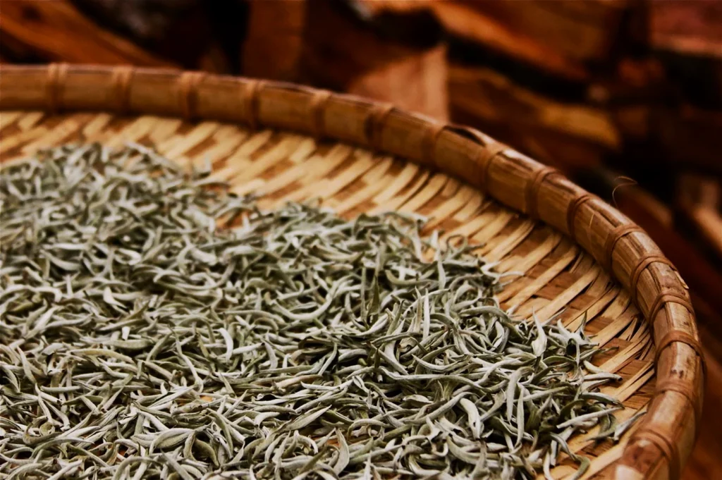 A round wicker tray holds a pile of pale green, needle-like tea leaves. The background is a textured brown, evoking a warm and rustic atmosphere.