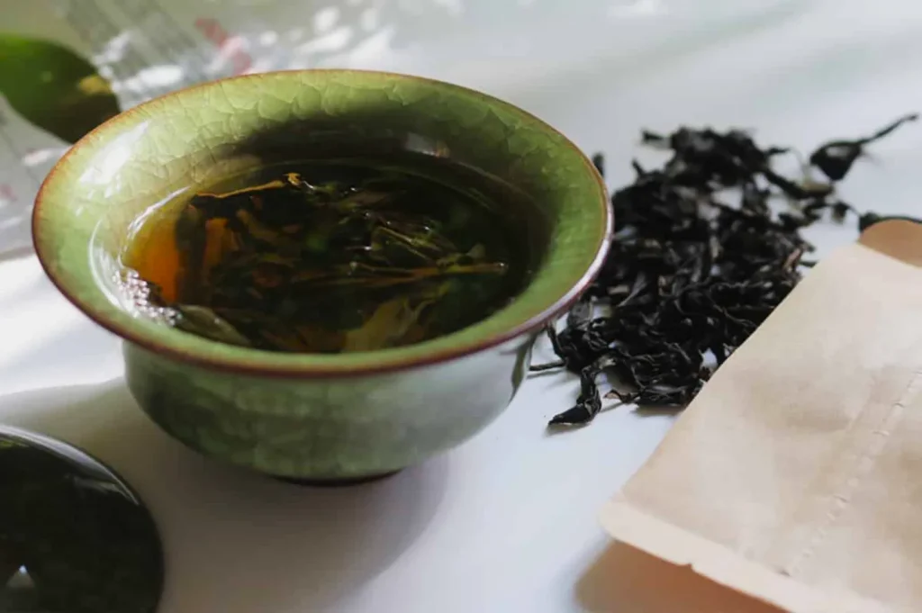 A green ceramic cup filled with steeping tea leaves sits on a white surface, alongside loose black tea leaves and an open brown teabag, evoking a tranquil tea-making scene.