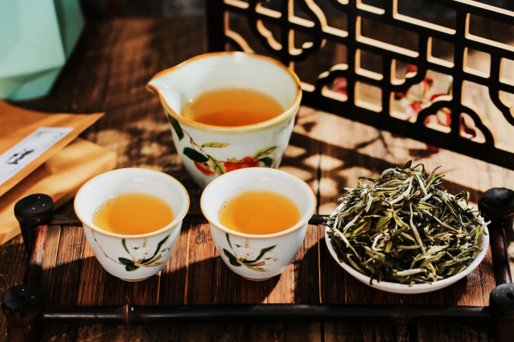 Tea setup with two small cups and a pitcher with light brown tea against a wooden backdrop. A bowl of dried tea leaves adds a rustic touch.