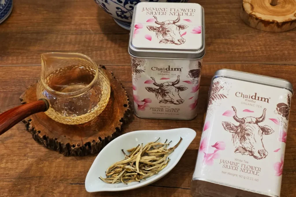 Wooden table with jasmine flower silver needle tea tins featuring cow illustrations. A glass teapot with brewed tea and loose tea leaves in a dish are nearby.