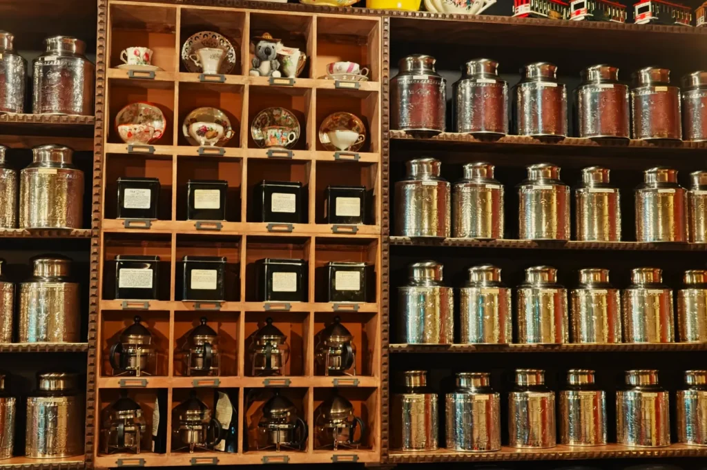 Wall with shelves of shiny metal tea canisters, ornate teapots, cups, and black boxes. Displays a cozy, vintage tea shop ambiance.