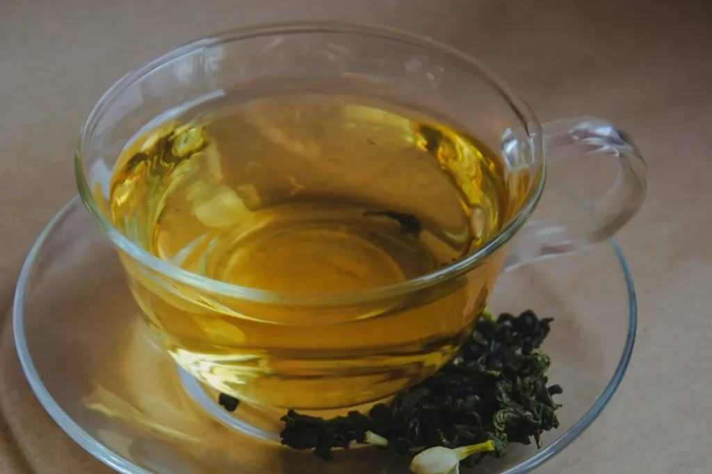A glass cup of golden green tea sits on a clear saucer, exuding warmth. Beside it, loose tea leaves add a natural, calming touch.