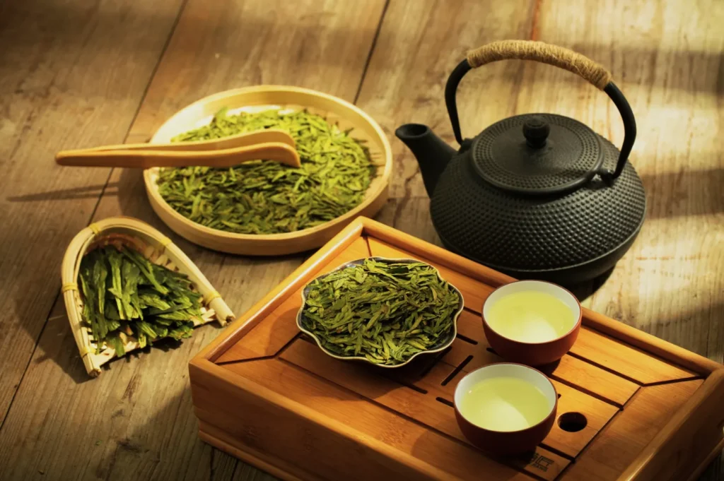 A serene tea setting with a black teapot, green tea leaves in a wooden dish, and two cups of tea on a bamboo tray, evoking a calm, traditional ambiance.