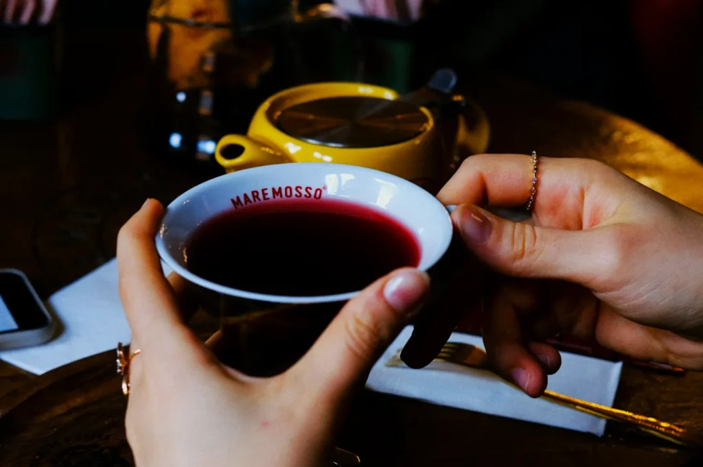 Hands holding a white cup filled with red tea labeled "MAREMOSSO" over a table. A yellow teapot and a phone are in the background, suggesting a cozy setting.
