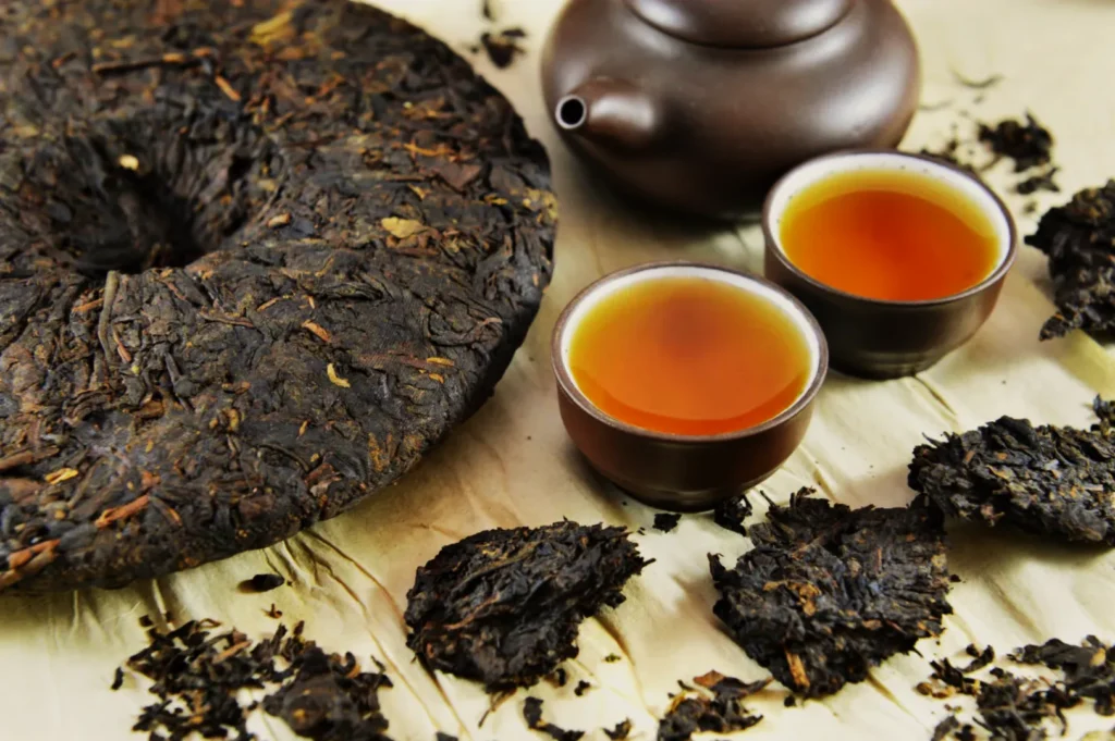 A round disc of compressed pu-erh tea is on a beige surface, with a brown teapot and two cups of amber tea nearby. Loose tea leaves are scattered around.