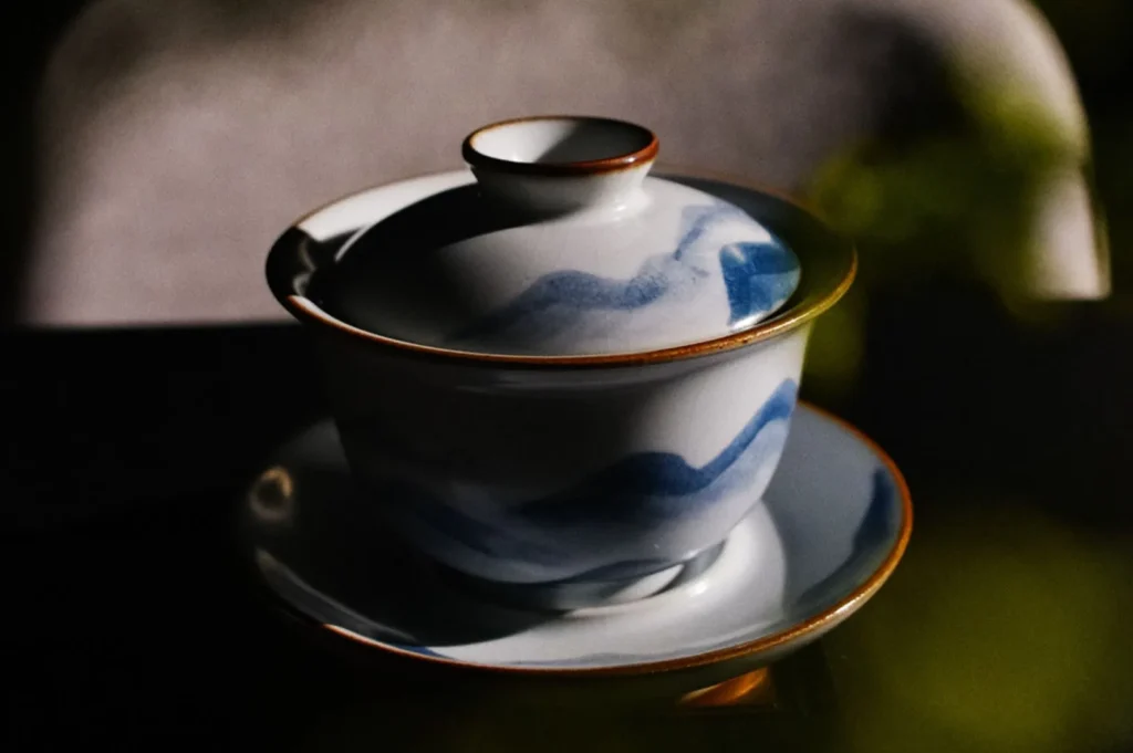 Ceramic teacup with lid on matching saucer, featuring blue wave patterns and brown edges, set against a softly blurred, dark background.