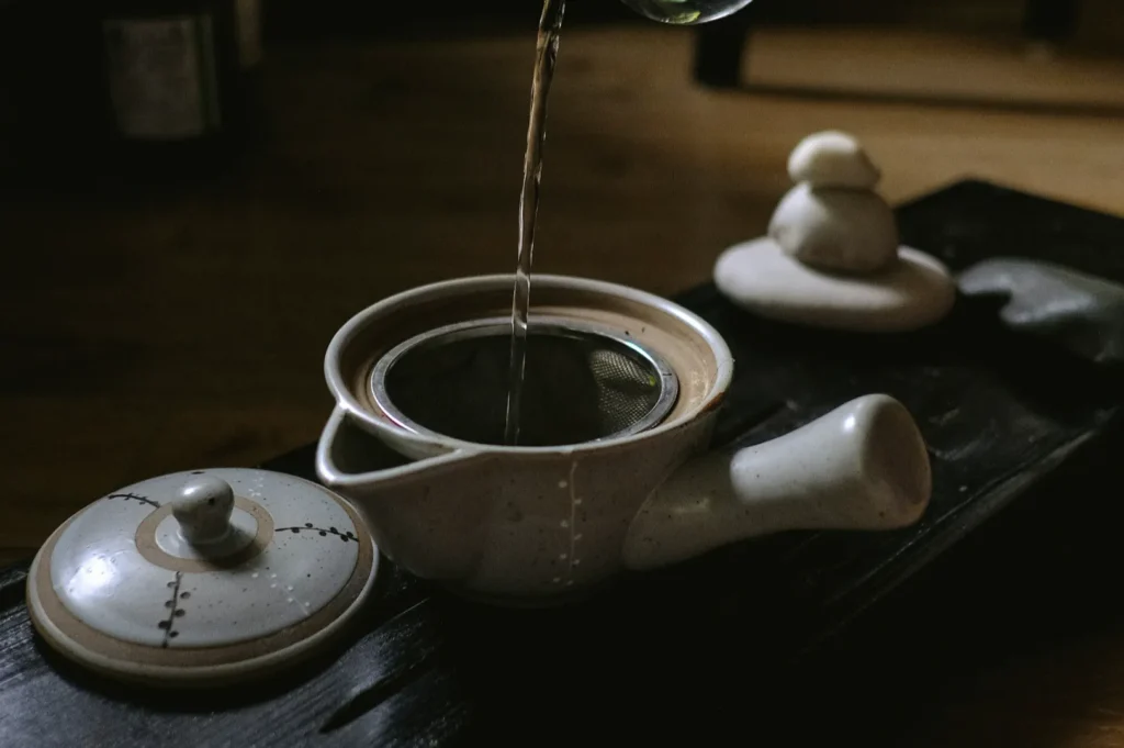 A cozy scene with tea being poured into a ceramic teapot through a strainer on a wooden tray. A stone decoration adds a tranquil touch. Dim lighting.
