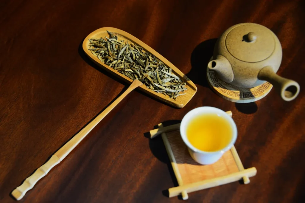 A wooden scoop filled with dried tea leaves, a ceramic teapot, and a steaming cup of tea rest on a wooden table, creating a cozy, inviting atmosphere.