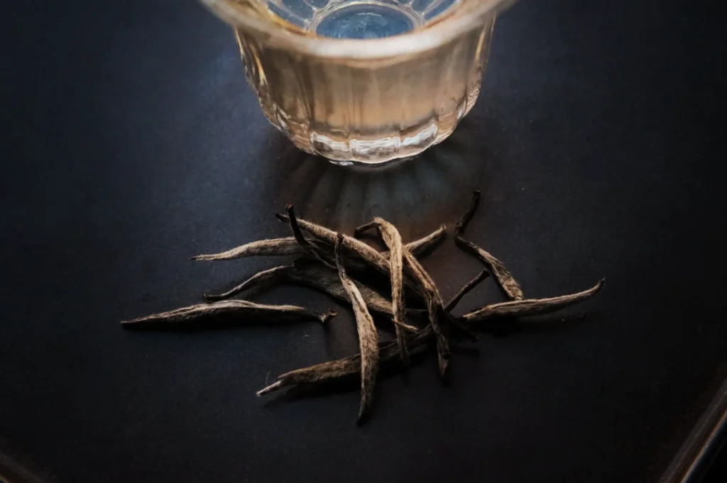 Glass teacup with light-colored tea next to a small pile of slender, dried tea leaves on a dark surface, conveying a calm, contemplative mood.